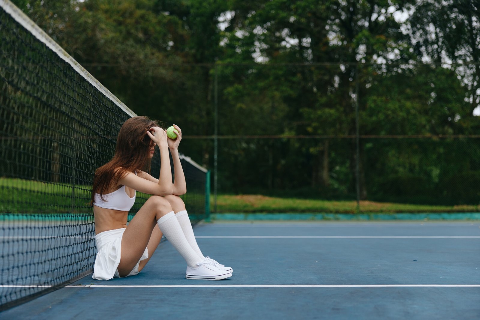 Young female tennis player in white sportswear sitting on a court, feeling frustrated with a tennis ball, surrounded by greenery, capturing the essence of sports emotion